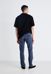 Zwarte korte mouwen shirt, blauwe spijkerbroek en zwarte sneakers. Witte stiksels op de jeans, gestructureerde shirtstof en standaard pasvorm.