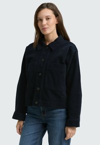 Vrouw in een donkerblauwe corduroy blousejas met grote voorzakken en blauwe spijkerbroek, staande tegen een effen achtergrond.