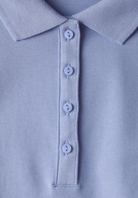Světle modrá polo košile s texturovanou látkou, která má klasický límec a čtyři odpovídající knoflíky podél zapínání.