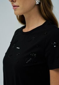 Mujer vestida con una camiseta negra con sutiles embellishments de perlas florales negras y un alfiler decorativo plateado en el pecho.