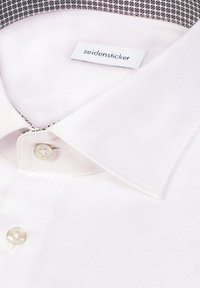 Weißes Hemd mit strukturierter Oberfläche, ausgestattet mit einem spitzen Kragen und einem kontrastierenden schwarzen und weißen gemusterten Innenkragen. Sichtbarer Knopfverschluss.