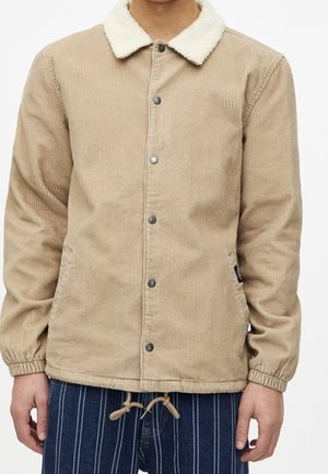 Veste en velours côtelé beige avec col en polaire blanc, boutons-pression, poignets élastiques, portée sur un pantalon bleu marine à rayures verticales blanches et cordon de serrage.