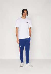 Balta kokvilnas T-krekls ar mazu melnu logotipu, kombinēts ar ziliem sporta biksēm, kurās ir sarkanas un baltas trīsstūra formas un potīšu manžetes.