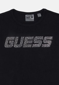 Juodas marškinėliai su žodžiu "GUESS", išsiuvinėtu sidabro spalvos blizgučiais. Apvalus iškirpimas ir minkšta medvilnė su lygiu tekstūru.