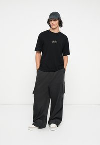 Musta crew neck -t-paita logolla, yhdistettynä löysiin harmaisiin cargo-housuihin ja kokonaisuutta täydentäviin valkoisiin tennareihin. Asusteina harmaa lippalakki.