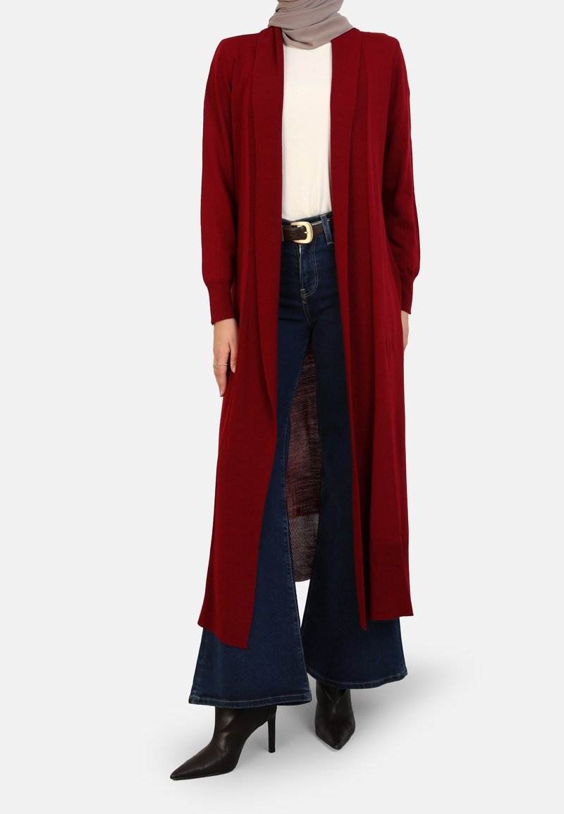 Femme portant un hijab beige, un long cardigan rouge ouvert, un haut blanc, un jean évasé foncé avec ceinture et des bottes noires à talons pointus.