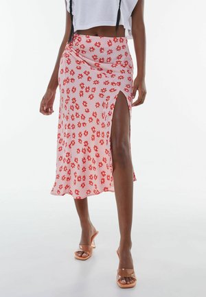 Vrouw draagt een roze midi-rok met rood bloemmotief en een hoge split tot op de dij, gecombineerd met transparante hoge hakken sandalen en een wit cropped topje.