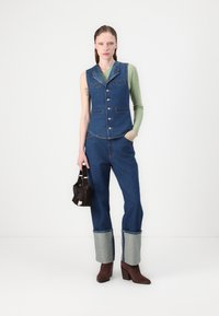 Jeans brezrokavnik z petimi gumbi, modrimi odtenki in sprednjimi žepi, v kombinaciji s širokimi modrimi jeans hlačami z svetlo modrimi manšetami. Rjave škornje s peto.