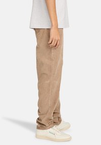 Hellbraune Cord-Hosen mit lockerer Passform, ausgestattet mit vertikal geripptem Textur, kombiniert mit weißen Sneakern und einem grauen T-Shirt.
