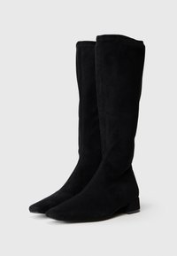 Botas de cano alto pretas, feitas de camurça, com bico fino e um pequeno salto bloco. Apresentam uma textura suave e costuras laterais discretas.