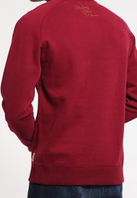 Tummanpunainen collegepaita pyöreällä pääntiellä, resorit hihansuissa ja helmassa, yläselässä pieni kulta tekstidetalji. Pehmeä, teksturoitu kangas.