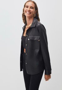 Giacca camicia nera in ecopelle con colletto, accessori in oro e tasca sul petto. Texture liscia e chiusure con bottoni lungo la parte anteriore.