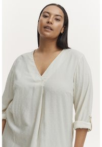 Blouse blanche légère avec un col en V, des manches trois-quarts et un motif texturé de petits points, présentant des poignets retroussés.