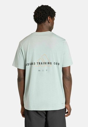 Mann mit kurzen lockigen Haaren, der ein hellgrünes Adidas Training Club T-Shirt und dunkle Shorts trägt, von hinten vor einem einfarbigen Hintergrund gezeigt.