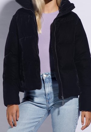 Personne portant une veste matelassée noire par-dessus un haut lavande et un jean bleu clair, debout devant un fond uni.