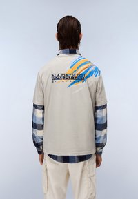 Beige langærmet skjorte med blå og orange grafisk tryk på ryggen; lagdelt over en blå ternet flannelskjorte; elfenbensfarvede bukser.