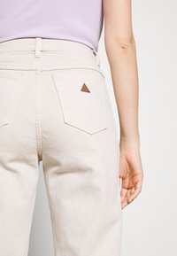 Beige denim pants with a high waist, featuring back pockets and a brown triangular leather patch. Smooth texture and casual design.