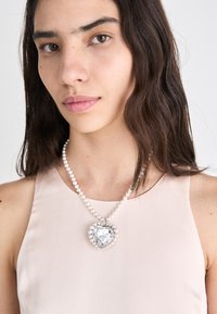 Pearl necklace with a large heart-shaped pendant, featuring a clear gem surrounded by small pearls, worn over a light pink top.