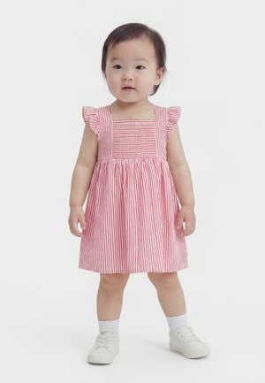 Νήπιο που στέκεται, φοράει ροζ και λευκό ριγέ φόρεμα με βολάν στα μανίκια, λευκές κάλτσες και λευκά παπούτσια σε απλό λευκό φόντο.