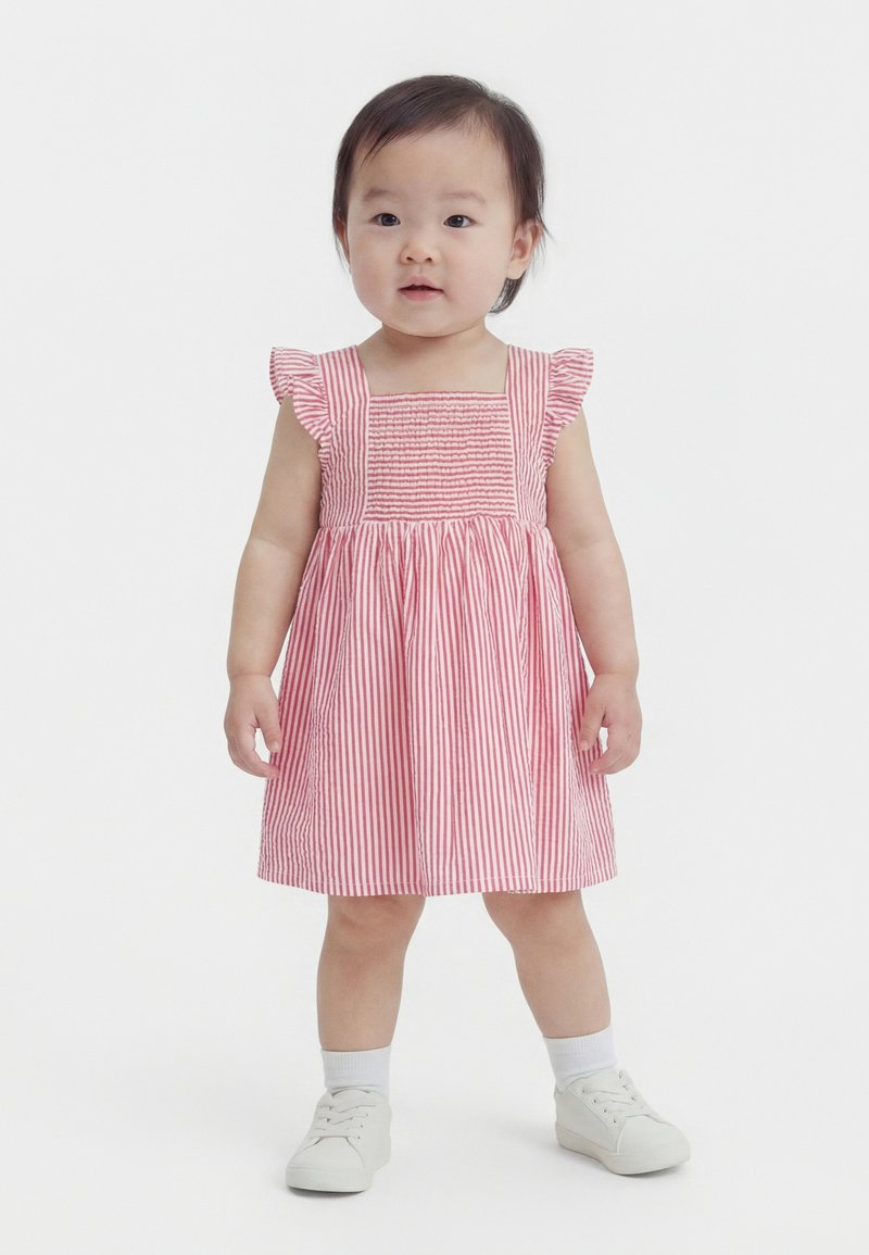 Copil mic în picioare, purtând o rochie cu dungi roz și albe, cu mâneci cu volane, șosete albe și pantofi albi, pe un fundal alb simplu.