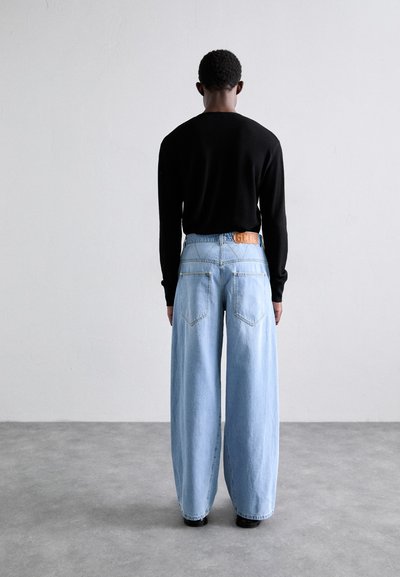 Camisa negra de manga larga combinada con jeans de mezclilla azul claro de pierna ancha. Los detalles notables incluyen grandes bolsillos traseros y un cinturón de cuero marrón con logo.