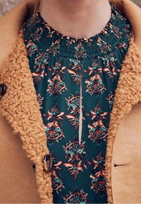 Vestido con estampado floral sobre fondo verde. Escote fruncido, finos pliegues, combinado con abrigo de textura beis y gran detalle de botón.