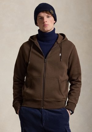 Jeune homme portant un sweat à capuche marron zippé sur un pull à col roulé bleu marine, un bonnet tricoté bleu marine et un pantalon bleu marine sur un fond uni.