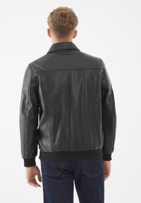 Mann mit kurzem blonden Haar, der eine schwarze Lederjacke und eine dunkelblaue Jeans trägt, von hinten vor einem weißen Hintergrund gesehen.