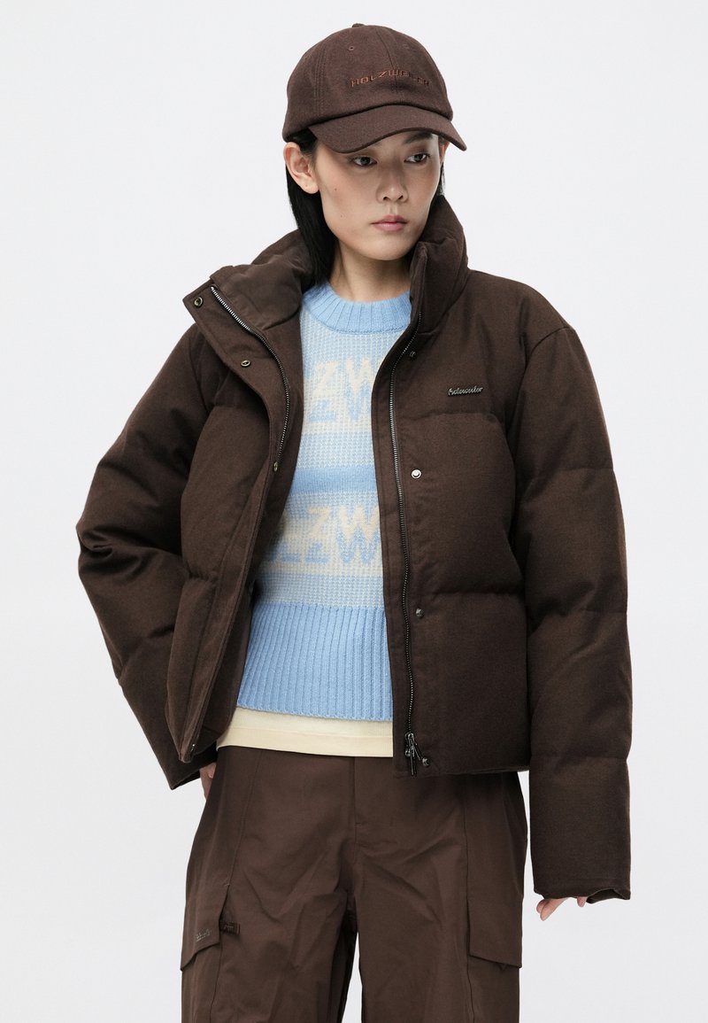 Brown puffer jacket with a high collar and zip closure. Worn over a light blue, patterned sweater. Accompanied by a matching cap.