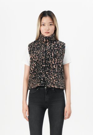 Femme portant un gilet sans manches en polaire à imprimé léopard sur un t-shirt blanc et un jean noir, se tenant contre un fond blanc uni.