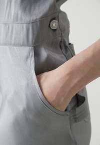 Γκρι παντελόνι από υφασμάτινη υφή, που διαθέτει τσέπη και μεταλλικό κουμπί διακοσμητικό. Ο σχεδιασμός είναι κομψός, με ορατές ραφές.