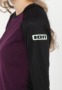 Langärmliges Sportoberteil in dunkellila und schwarz mit Netzstruktur und einem hellgrünen Logo auf dem linken Ärmel.