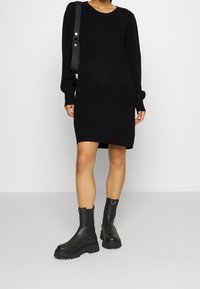 Robe noire en maille avec manches longues, poignets côtelés et coupe décontractée. Associée à des bottines noires en cuir avec une semelle épaisse.