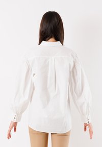 Blusa bianca con maniche a sbuffo, polsini abbottonati e una piega sul retro. Il tessuto appare liscio e simile al cotone, offrendo una vestibilità strutturata.