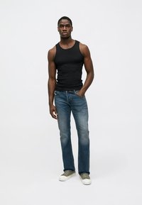 Top de algodão preto, jeans azul de lavagem ligeira, ténis cinzentos. Modelo em pé contra um fundo branco, com os braços relaxados ao lado.