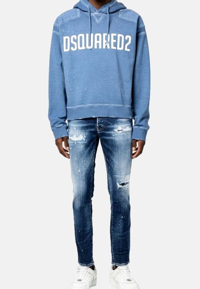 Blauer Kapuzenpulli mit weißem "DSQUARED2" Logo, kombiniert mit ausgewaschenen blauen Jeans mit Löchern. Weiße Sneakers vervollständigen das Outfit.