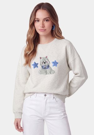 Giovane donna che indossa una felpa beige chiaro con un cane bianco ricamato e stelle blu, abbinata a pantaloni bianchi, in piedi davanti a uno sfondo semplice.