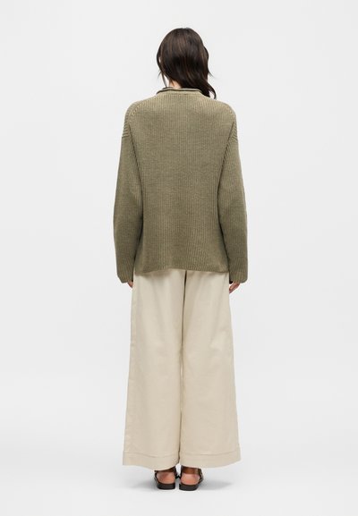 Femme de dos portant un pull en maille vert olive, un pantalon large beige et des sandales noires sur un fond gris uni.
