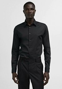 Cămașă neagră cu nasturi, cu guler ascuțit, mâneci lungi și un material neted și structurat, asociată cu pantaloni eleganți asortați.