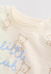 Chemise pour bébé couleur crème avec des contours d'ourson marron clair et le mot « little » écrit en cursive bleue près du col.