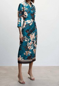 Teal bloemen wikkeljurk van satijn met 3/4 mouwen, gekenmerkt door perzikkleurige en zwarte bloemen, een gestrikt middel en een effen bruine zoom met strepen.