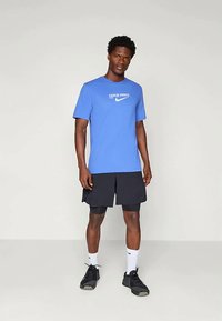 Mann som står iført blå Nike Pro t-skjorte, svarte lagdelte shorts med kompresjon, hvite Nike sokker og svarte treningssko.