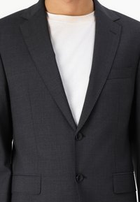 Mörkgrå blazer i strukturerad tyg, med snibb-lapel, två knappar och bröstficka. Bärs över en vit skjorta.