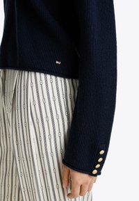 Granatowy sweter z prążkowanego materiału z dodatkami złotych guzików na mankietach, zestawiony z kremowo-granatowymi spodniami w paski z prostokątnymi wzorami.