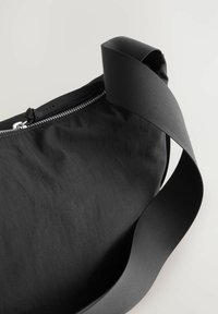Bolsa de tela negra con una textura suave, que cuenta con un cierre de cremallera y una amplia correa negra acanalada para un fácil transporte.