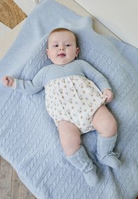 Manta de punto azul claro con un patrón texturizado, un bebé vestido con una camiseta azul y un mono blanco con pequeños patrones coloridos, y botines de punto azules.