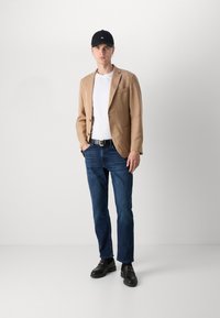 Blazer marrone chiaro sopra una t-shirt bianca, jeans blu e scarpe slip-on nere. Un berretto nero e un orecchino a bottone completano il look.