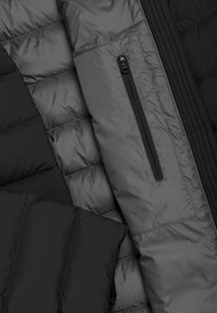 Črn napihnjen jopič z kvadratnim vzorcem, gladko teksturo in žepom z zadrgo na rokavu. Lahka in izolirana tkanina.