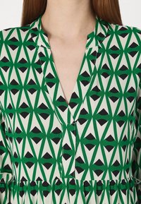 Vihreä ja valkoinen kuvioitu mekko, jossa on geometrinen muotoilu mustilla yksityiskohdilla, kauluksessa on halkio ja hameosa on pleated.