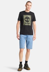Camiseta de algodón negra de manga corta con un gráfico del logo de Timberland en camuflaje verde. Combinada con pantalones cortos de mezclilla azul y zapatos de senderismo negros.